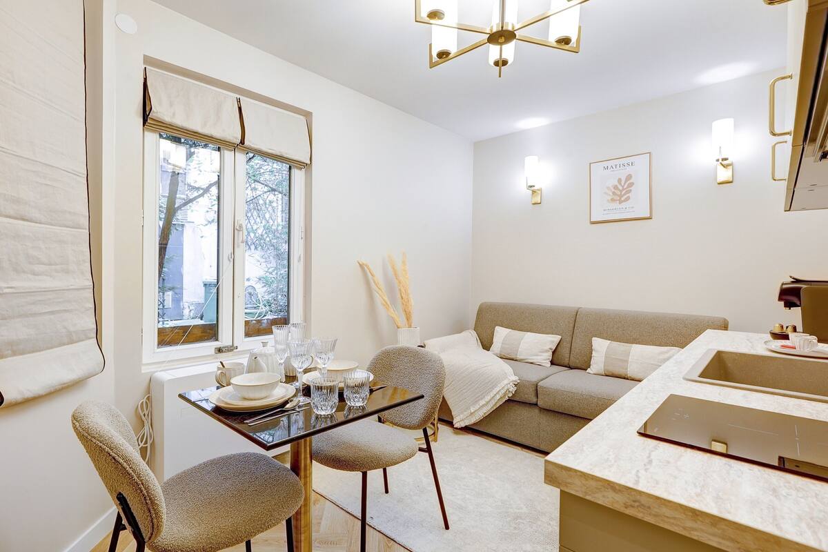 A modern living room with a sofa, a dining table with chairs, a kitchen area, and a window with blinds.