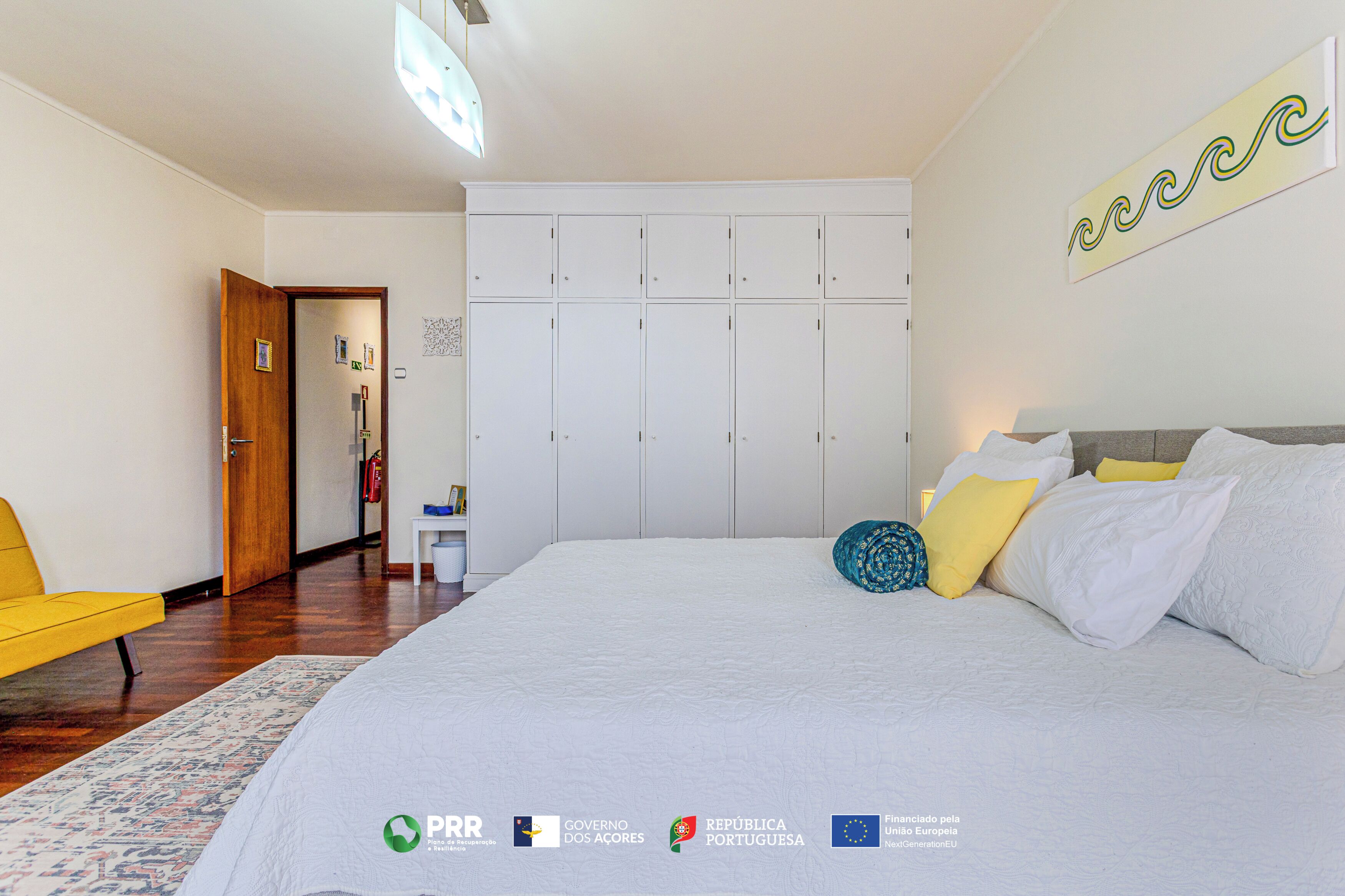 A bedroom with a bed, a wardrobe, a chair, and a wall with a decorative piece.
