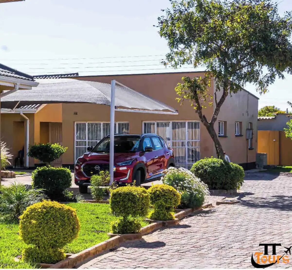 Un SUV rouge était garé dans une allée menant à une maison, devant une terrasse couverte, le tout entouré d’arbustes bien entretenus et d’un arbre.