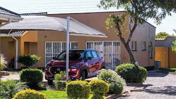 Un SUV rouge était garé dans une allée menant à une maison, devant une terrasse couverte, le tout entouré d’arbustes bien entretenus et d’un arbre.