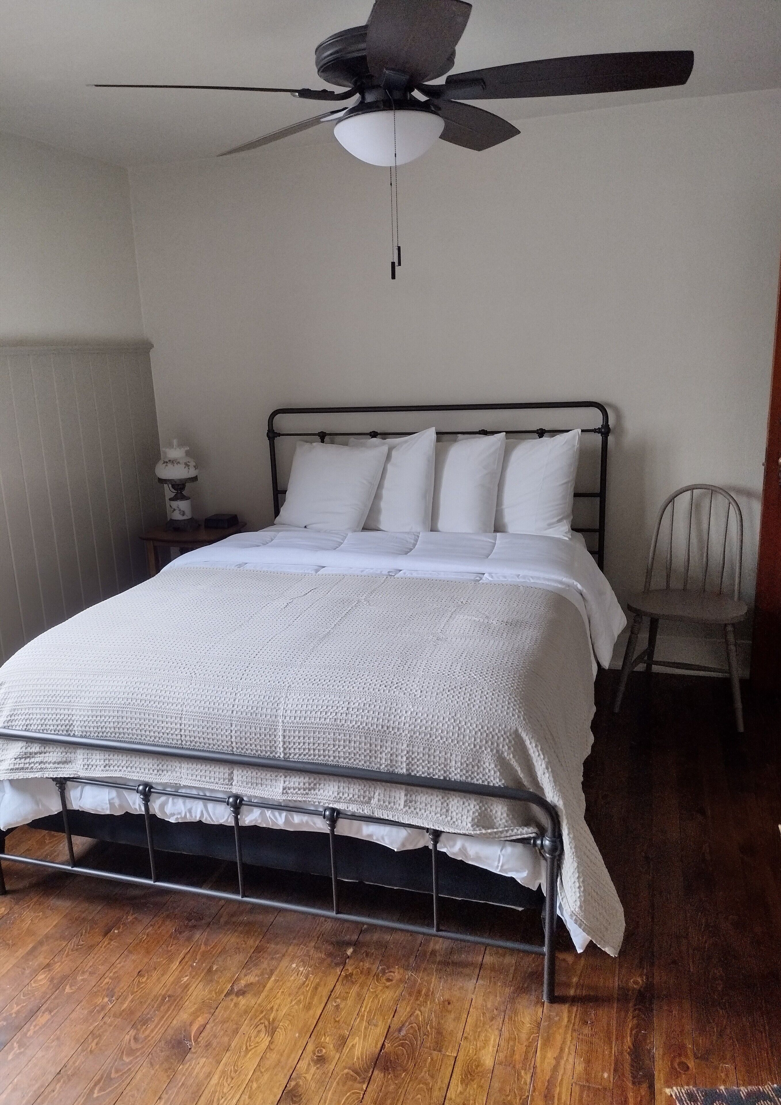 Quarto com estrutura de cama de metal, roupa de cama branca, ventoinha no teto, mesa de cabeceira com abajur e uma cadeira.