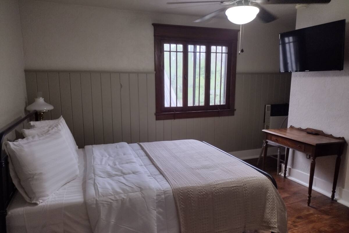 Ein Schlafzimmer mit Bett, Schreibtisch, Stuhl, Fenster und Fernseher an der Wand.