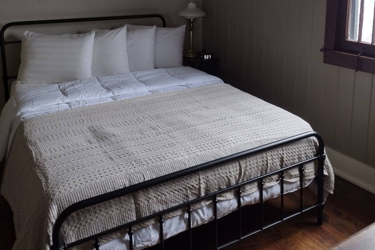 Ein Schlafzimmer mit einem Bett, einem Deckenventilator, einem Nachttisch, einer Lampe und einem Fenster.