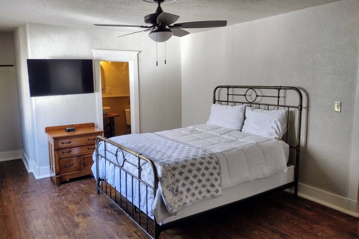 Ein Schlafzimmer mit einem Metallbettgestell, einem Deckenventilator, einer hölzernen Kommode und einem Fernseher an der Wand.