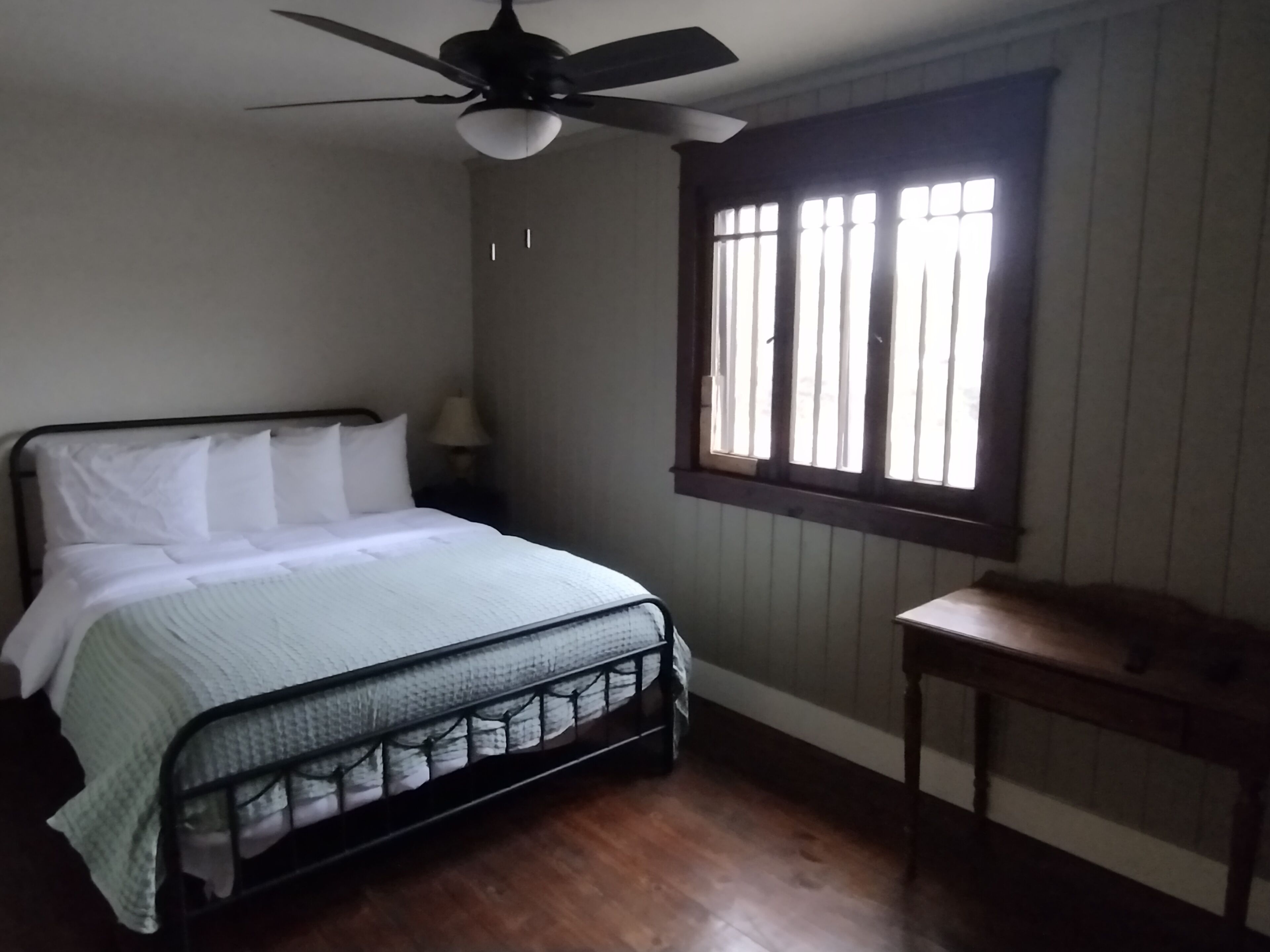 Un dormitorio con cama, ventilador de techo, ventana con persianas y una mesita.