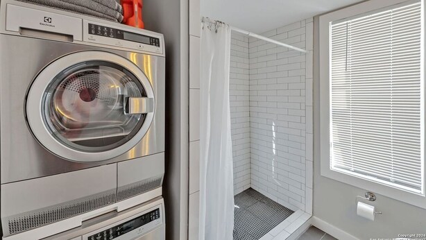 Ein kleiner Waschraum mit einer Waschmaschine mit Frontlader, einer Dusche mit weißer Duschvorhang und einem Fenster mit Jalousien.