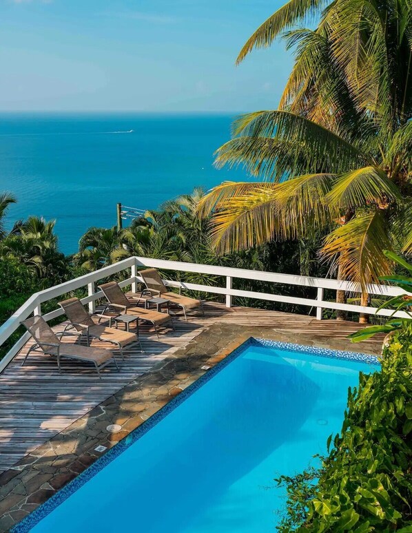 Une piscine dotée d’une terrasse en bois et de chaises longues, entourée de palmiers et offrant une vue sur l’océan.