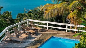 A pool with a wooden deck and lounge chairs, surrounded by palm trees and a view of the ocean. - Amazing mountain top villa! (Castries)