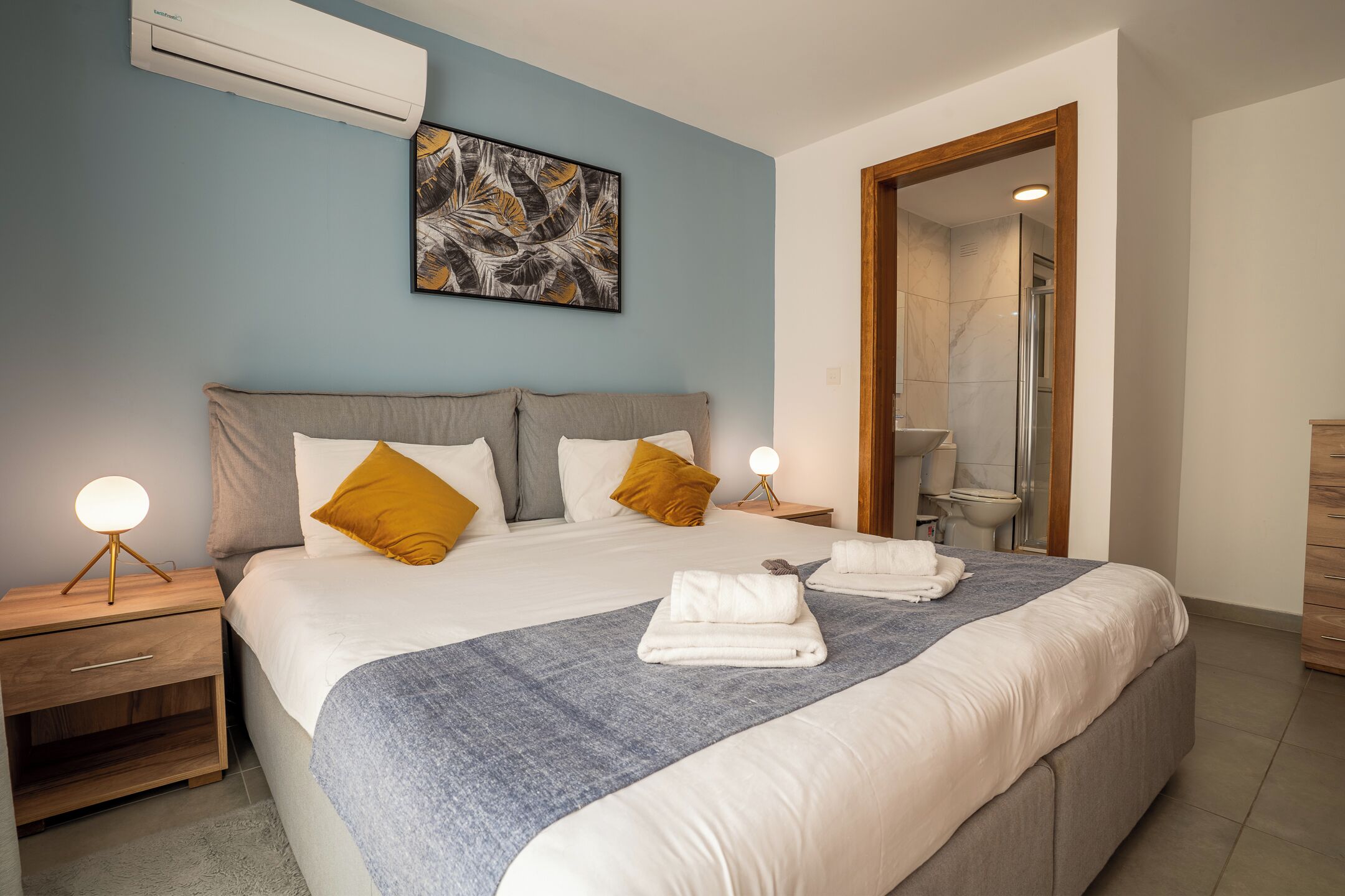 A neatly made bed with a blue blanket, two bedside tables with lamps, a wall-mounted air conditioner, and a bathroom visible through an open door.