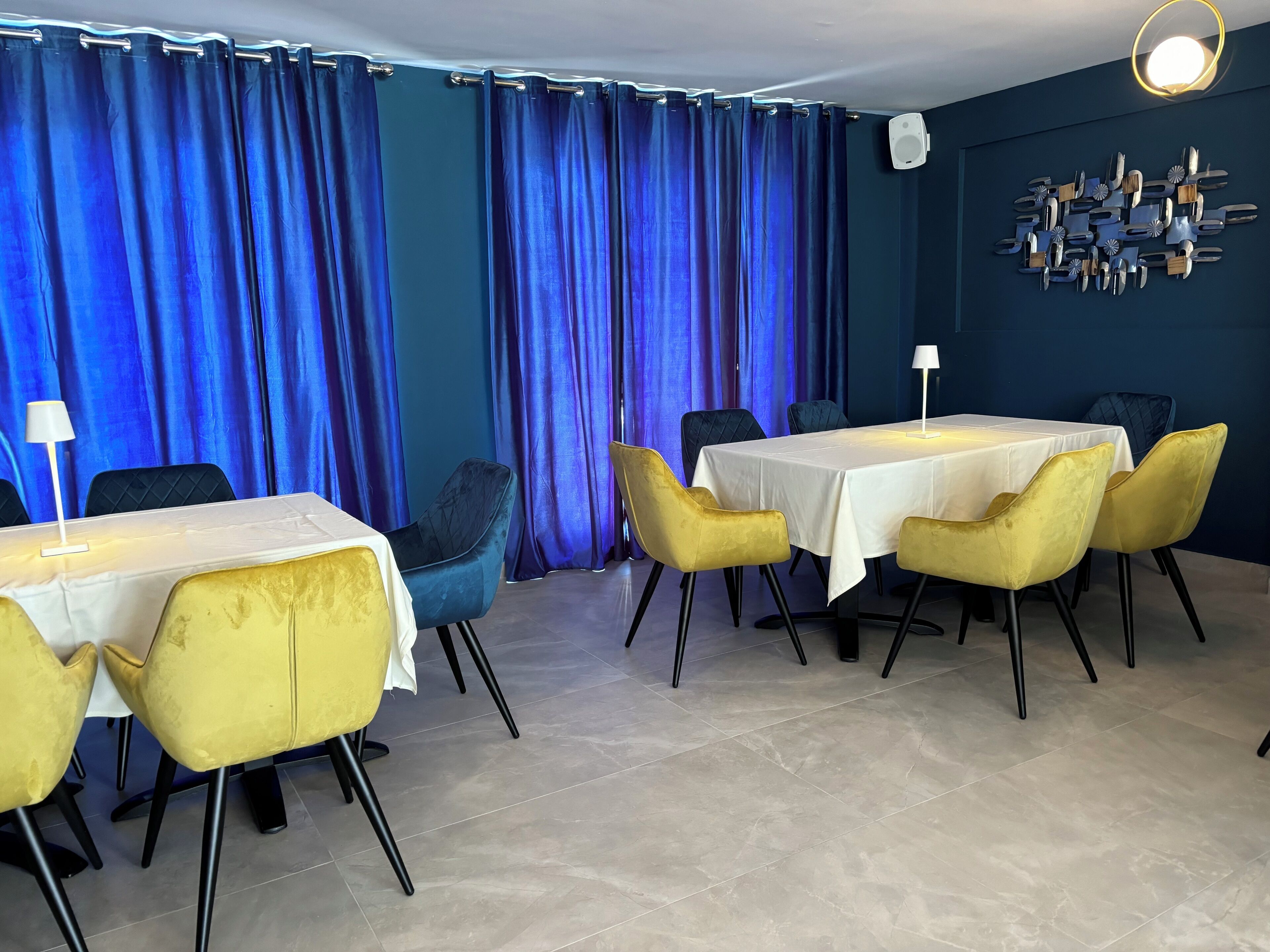 A dining area with tables and chairs, blue curtains, and a wall-mounted light fixture.
