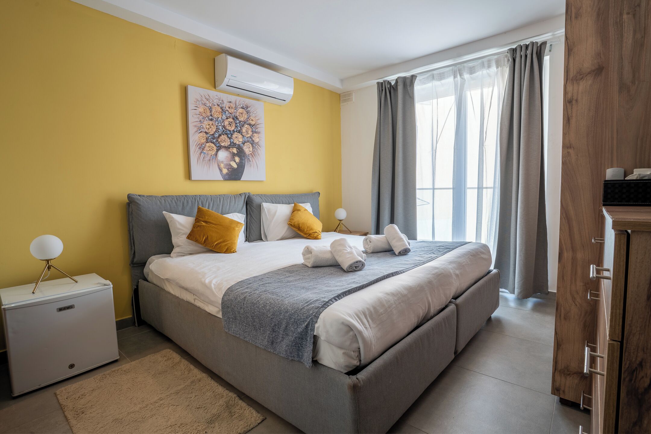 Un dormitorio con cama, mesitas de noche, aire acondicionado, una ventana con cortinas y un armario de madera.
