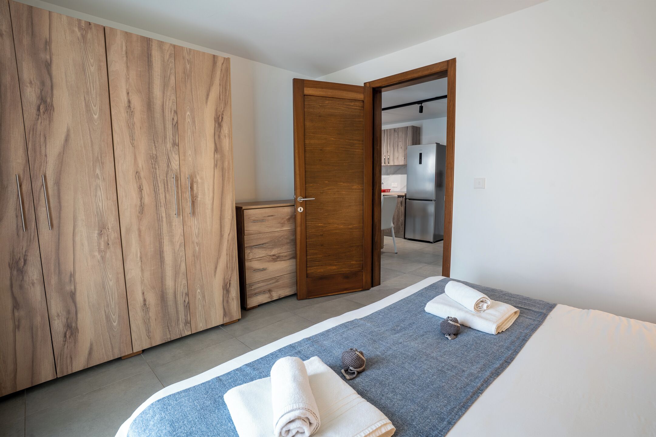 Een hotelkamer met een grote inbouwkast, een bed met een blauwe deken, en een kleine kitchenette die door een open deur zichtbaar is.