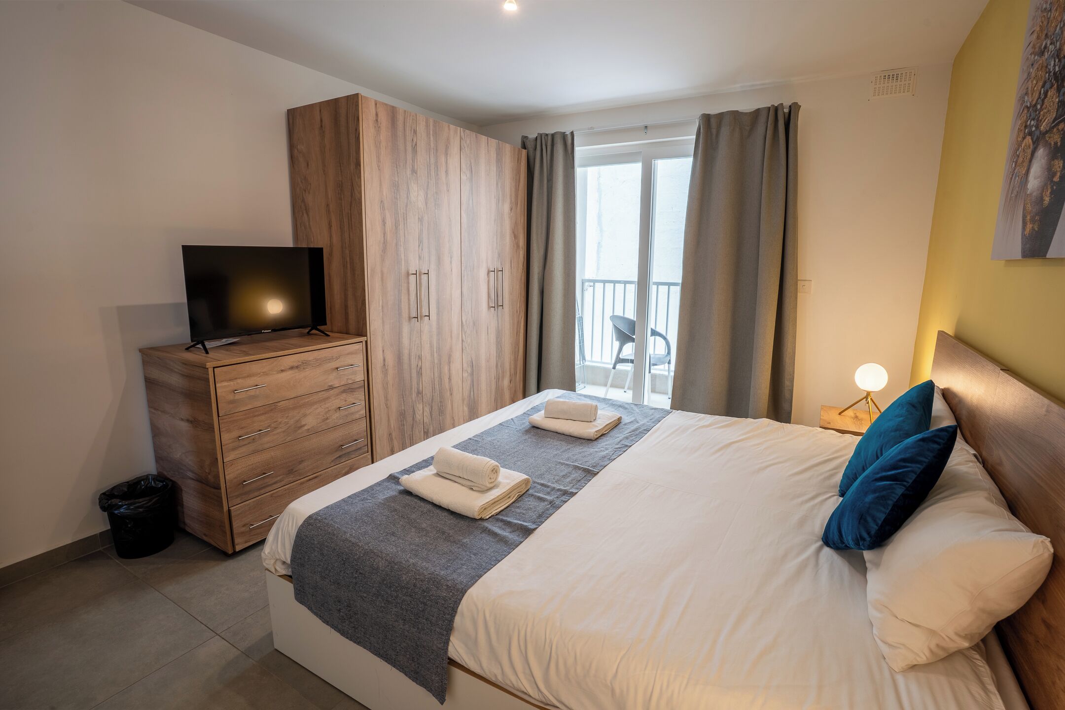 A hotel room with a bed, a TV, a wooden wardrobe, a balcony with curtains, and a nightstand with a lamp.