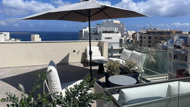 Terraza en la azotea con una sombrilla grande, mobiliario exterior y vistas al mar y edificios.