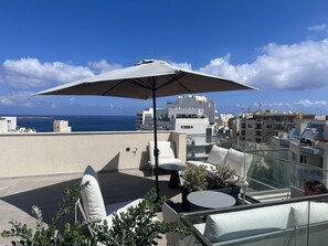 A rooftop terrace with a large umbrella, outdoor seating, and a view of the sea and buildings. - StiLLel Retreat (St. Paul's Bay)
