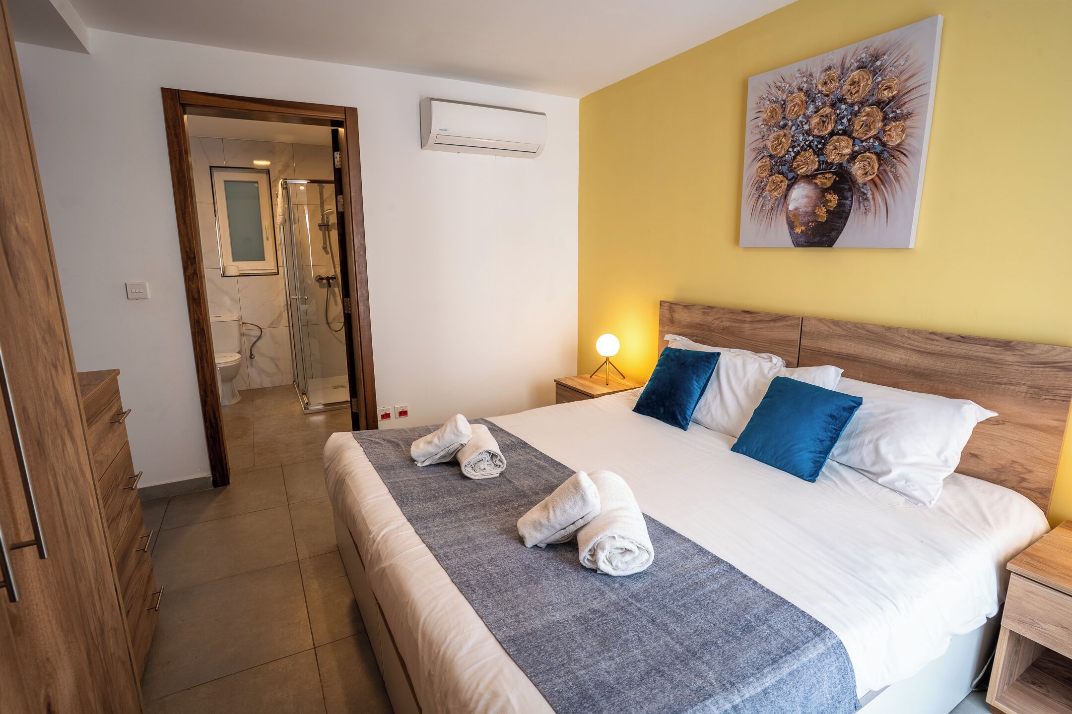 Una habitación de hotel con una cama, una mesita de noche, un espejo, un equipo de aire acondicionado y un cuadro en la pared.