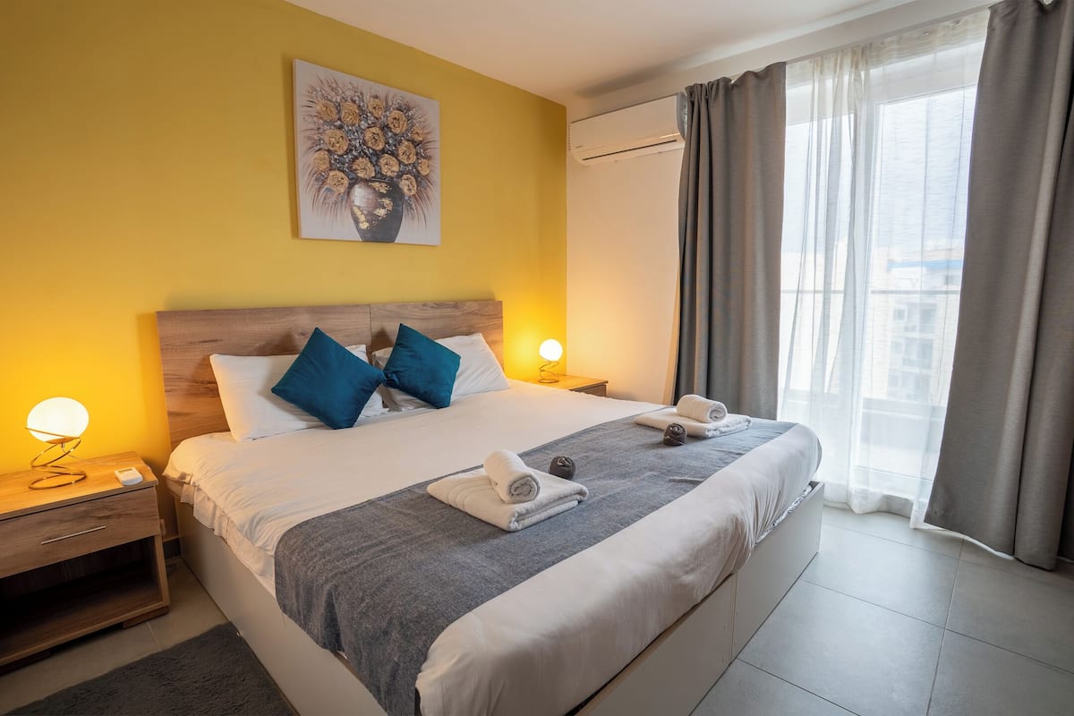Una habitación de hotel con una cama, lámparas de noche, un cuadro y un ventanal con cortinas.