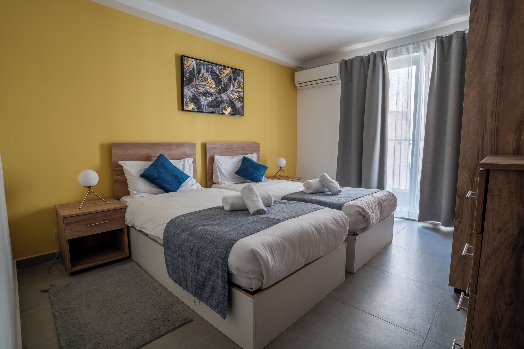 A hotel room with two beds, a nightstand, a lamp, a painting, and a view of the outdoors.