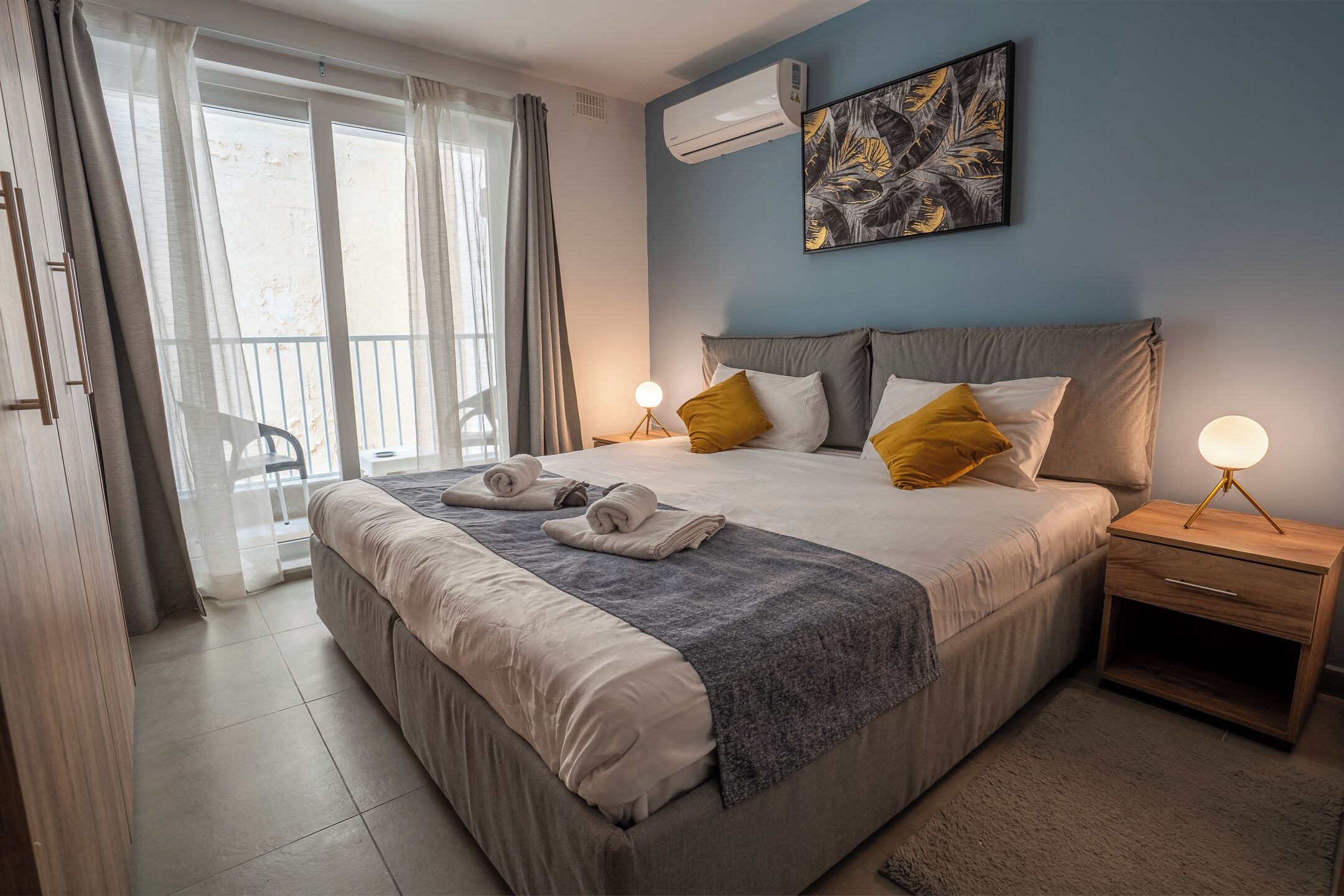 A bedroom with a bed, a nightstand, a wall-mounted air conditioner, a balcony with a chair, and a painting.
