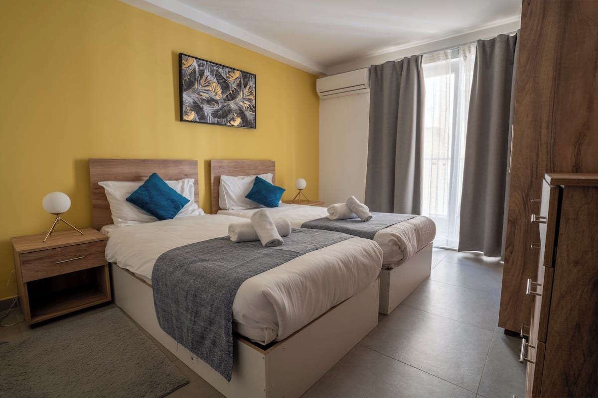 Habitación de hotel con dos camas, una mesita de noche con lámpara, un cuadro en la pared y vistas al exterior a través de la ventana.