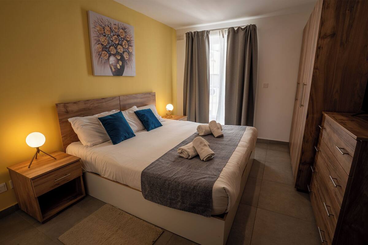 Un dormitorio con cama, mesitas de noche, cómoda, un cuadro y una ventana con cortinas.