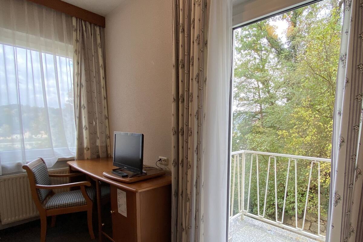 Habitación de hotel con escritorio de madera, una silla, televisor y balcón con vista a árboles.