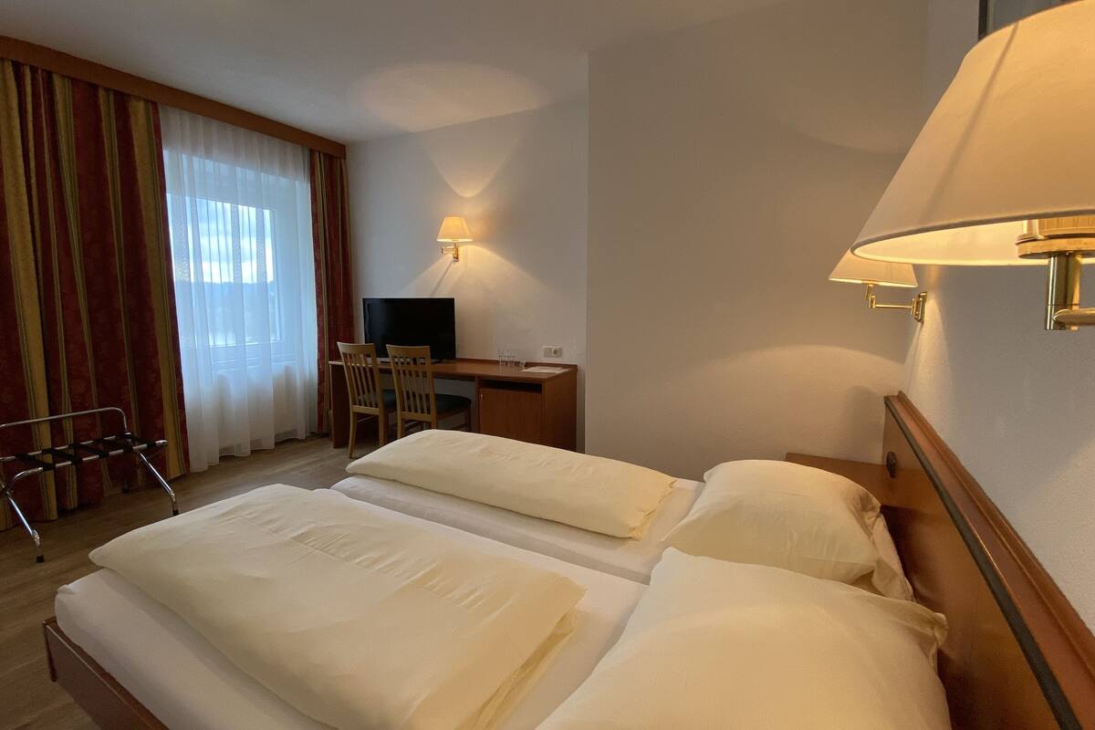 Habitación de hotel con dos camas, un escritorio con silla, televisor y ventana con cortinas.