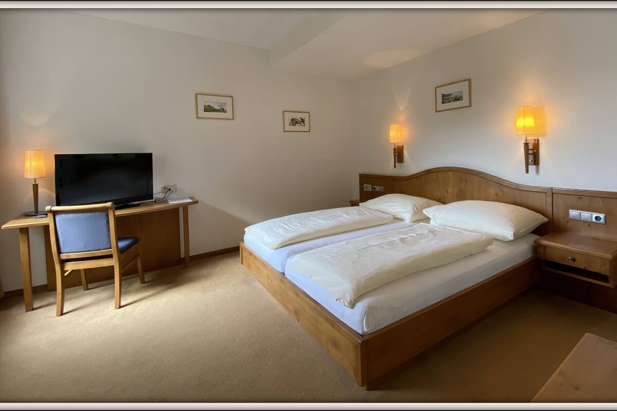 Una habitación de hotel con una cama de madera, un escritorio con televisor y dos lámparas fijadas a la pared.