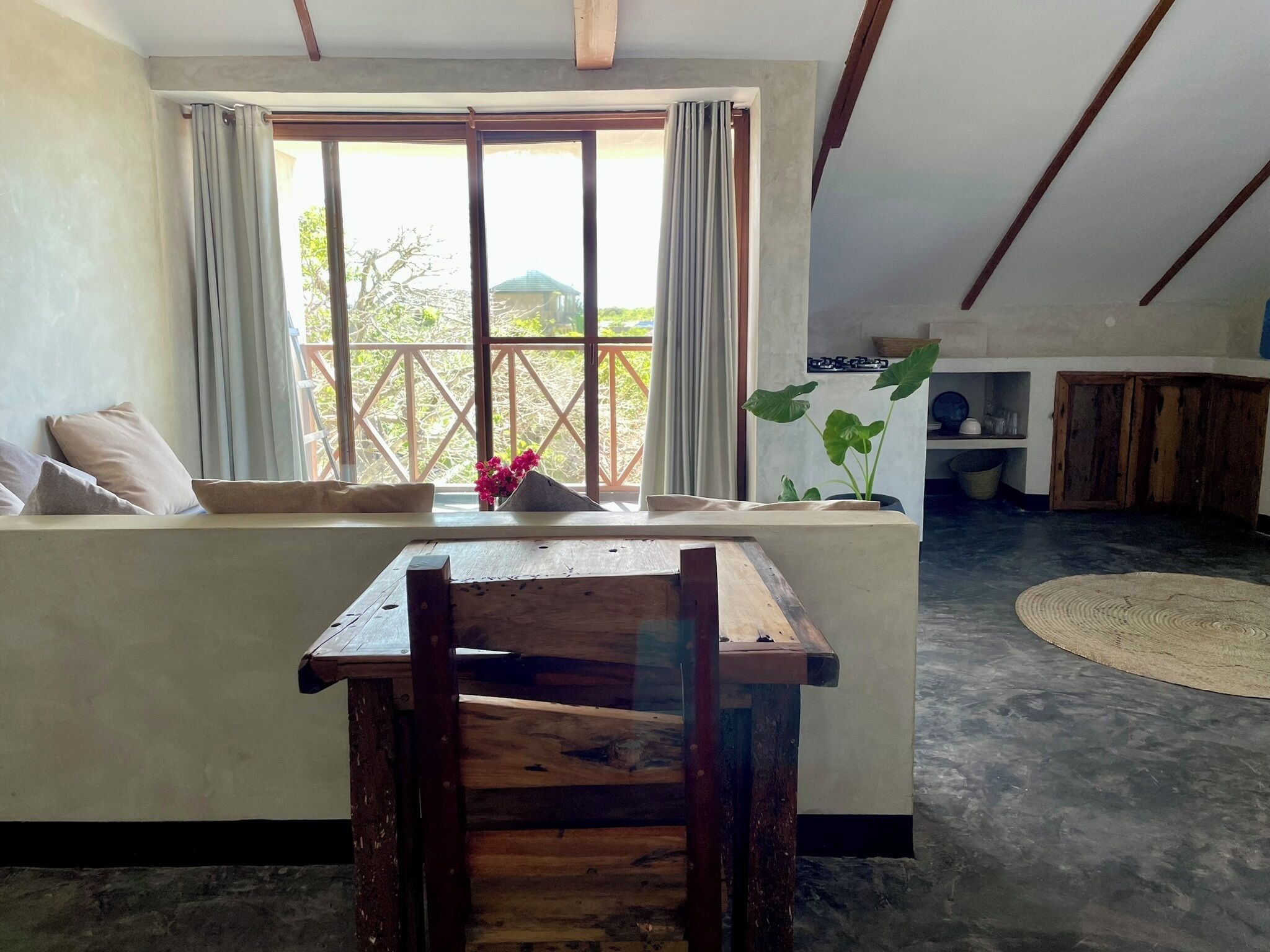 A room with a wooden table and chairs, a sofa, a window with curtains, and a view of greenery outside.