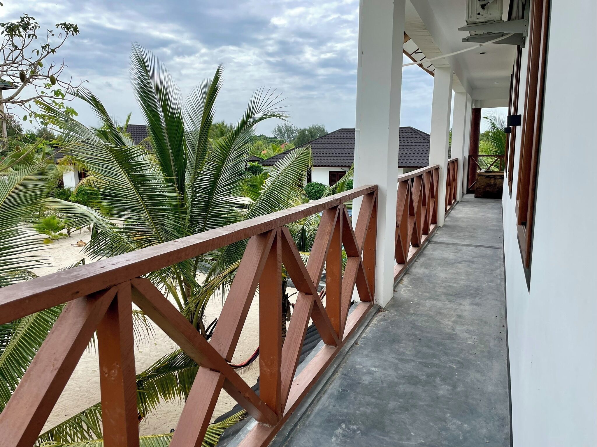 A covered balcony with a wooden railing, overlooking a sandy area with palm trees and a building with a tiled roof.