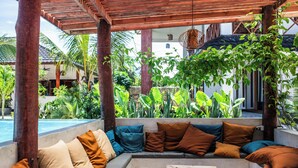 Een overdekt terras met een zwembad, omgeven door weelderige begroeiing en een houten pergola.