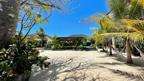 Een tropisch resort met een zandkoer, palmbomen en een gebouw met rieten dak.