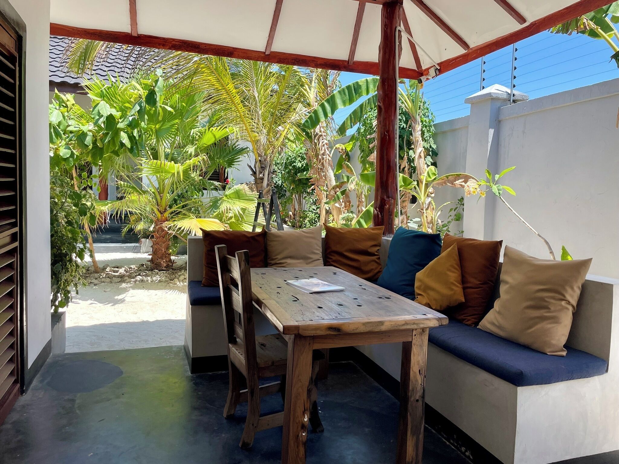 Un espacio de estar techado al aire libre con una mesa de madera y bancos, rodeado de exuberante vegetación y un muro blanco.