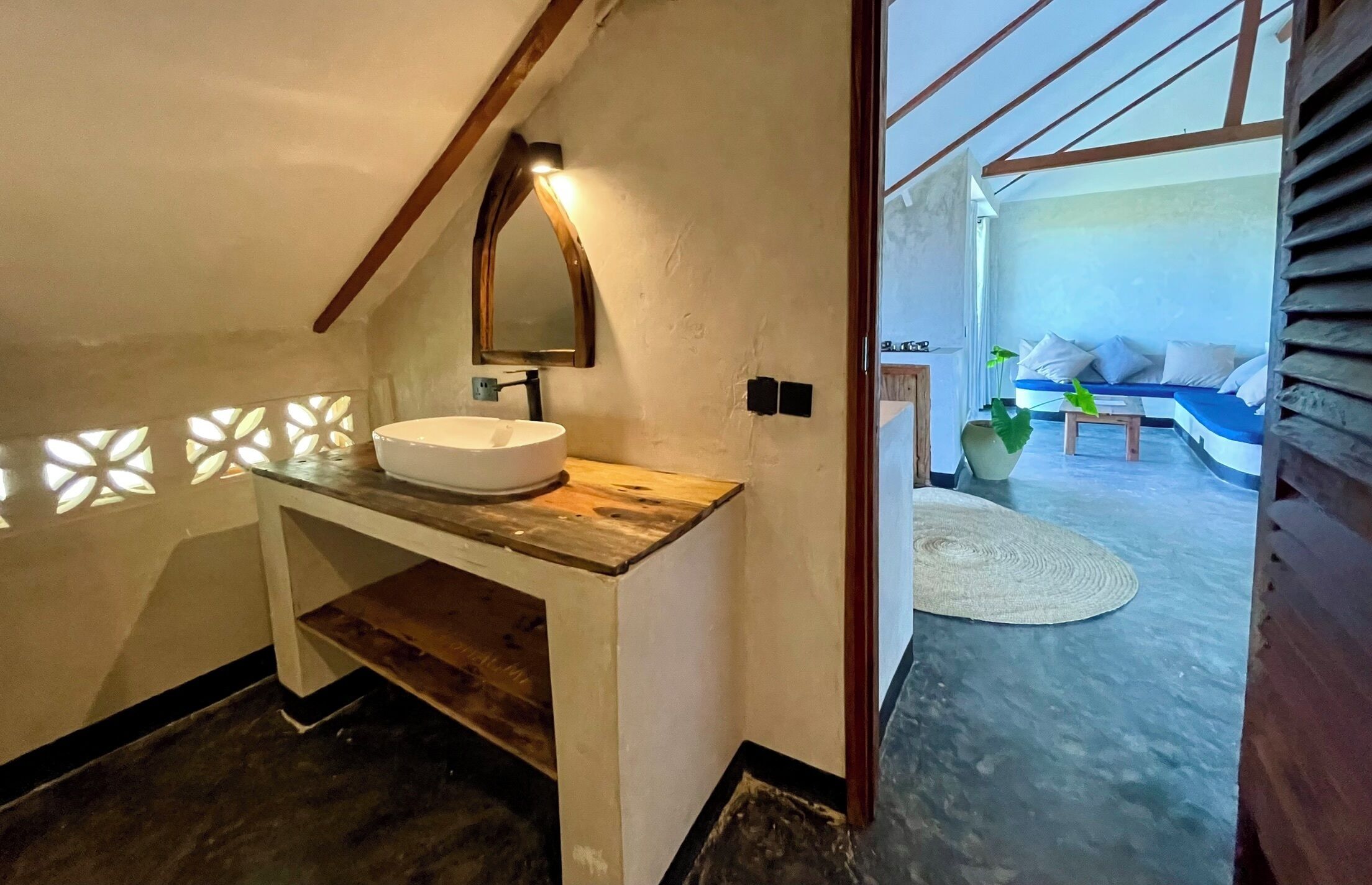 A bathroom with a wooden vanity, a white sink, and a mirror. There is a small table with a plant and a white rug in the adjacent living area.