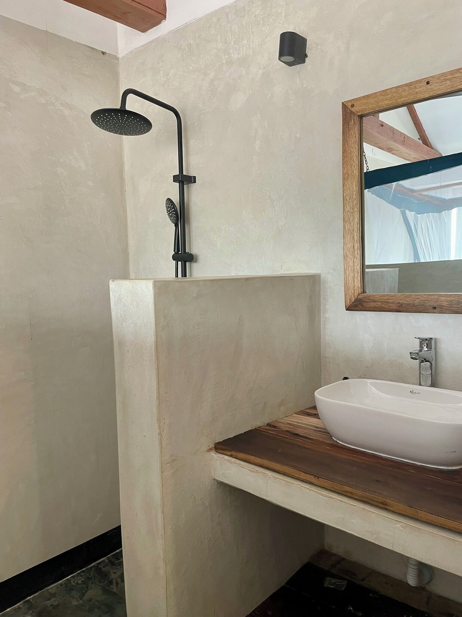 A bathroom with a white sink, a wooden vanity, a black showerhead, and a mirror with a wooden frame.