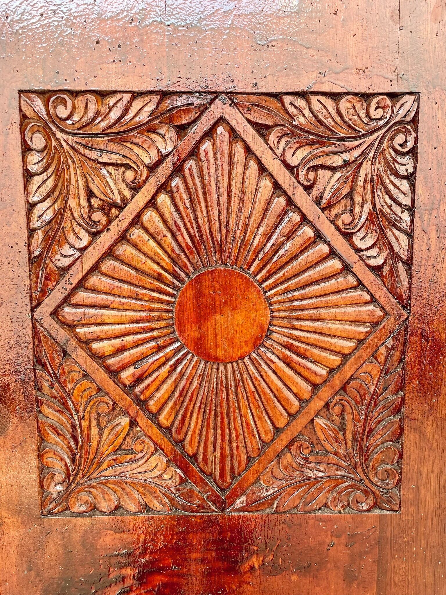 Un panneau en bois sculpté avec de nombreux détails, orné d’un motif central représentant un soleil, entouré de motifs géométriques et de motifs évoquant des feuilles.