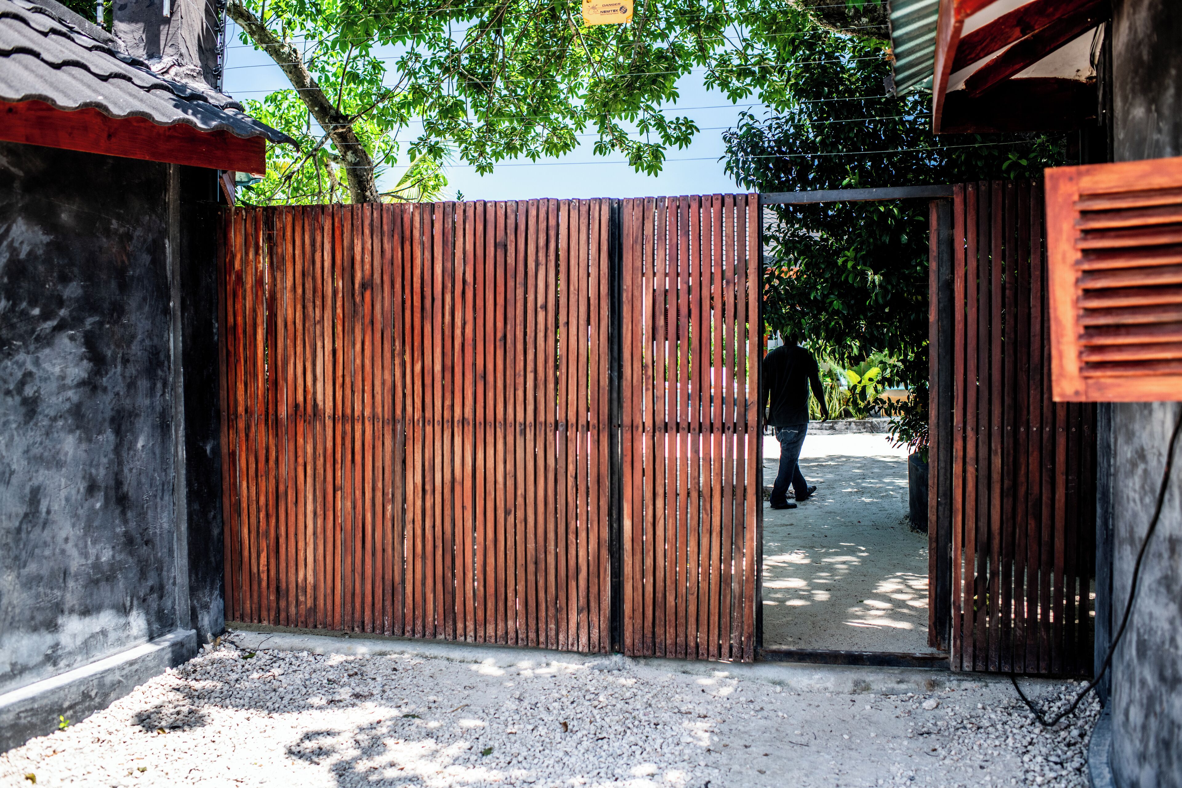 A gated entrance with a wooden fence, a concrete pathway, and a building with a corrugated metal roof.
