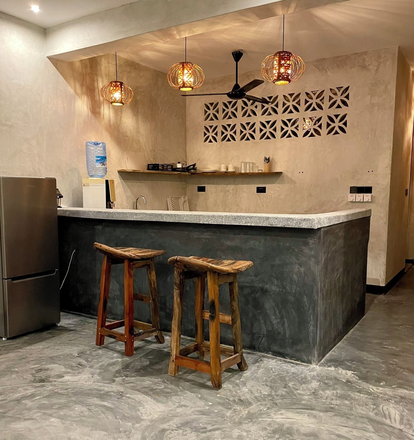 A modern kitchen with a central island, two wooden stools, pendant lights, a ceiling fan, and a decorative wall panel.