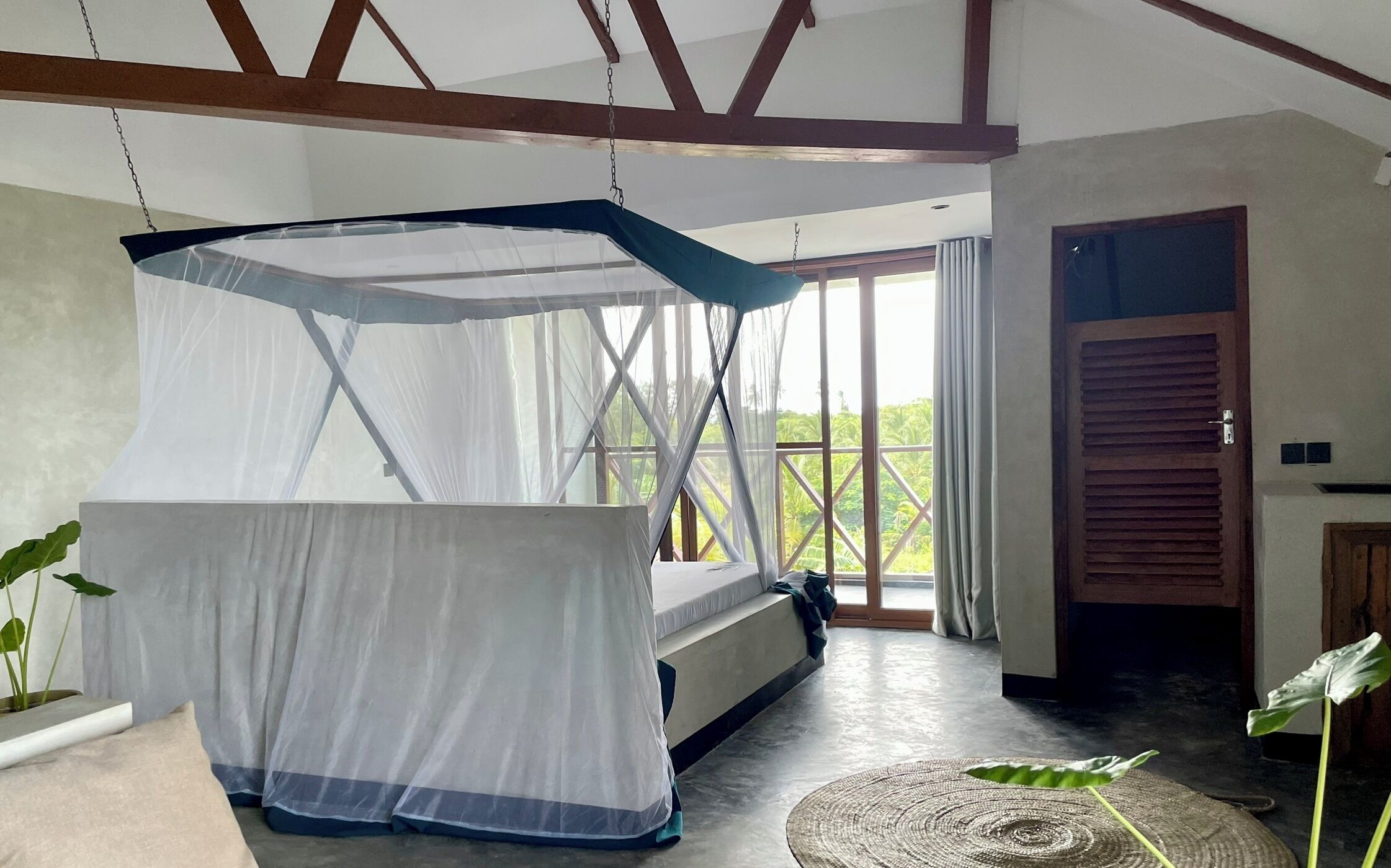 A room with a bed under a mosquito net, a large window with a view of greenery, and a door with a wooden shutter.