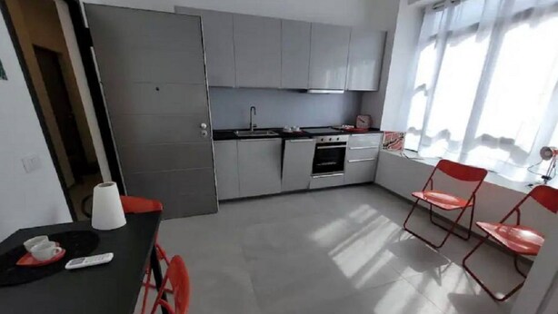 Une cuisine moderne avec des meubles gris, une cuisinière et un évier. On y trouve deux chaises rouges et une table sur laquelle sont posés une tasse et une télécommande.