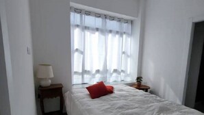 Une chambre à coucher avec un lit, une fenêtre ornée de rideaux légers, une table de chevet avec une lampe et une petite table sur laquelle est posée une plante.