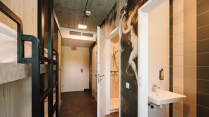 Un couloir étroit, avec des lits superposés sur le côté gauche, une salle de bain avec douche et lavabo sur le côté droit, et une porte donnant sur une autre pièce.