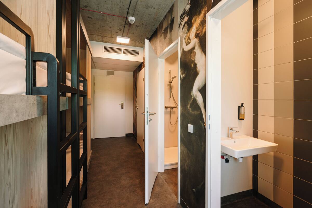 Ein schmaler Gang mit Etagenbetten links, einem Badezimmer mit Dusche und Waschbecken rechts sowie einer Tür zu einem weiteren Raum.