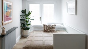 Ein heller, weißer Raum mit einem großen Fenster, einer Pflanze, einem Bett mit gemustertem Kissen und gerahmten Kunstwerken an den Wänden.