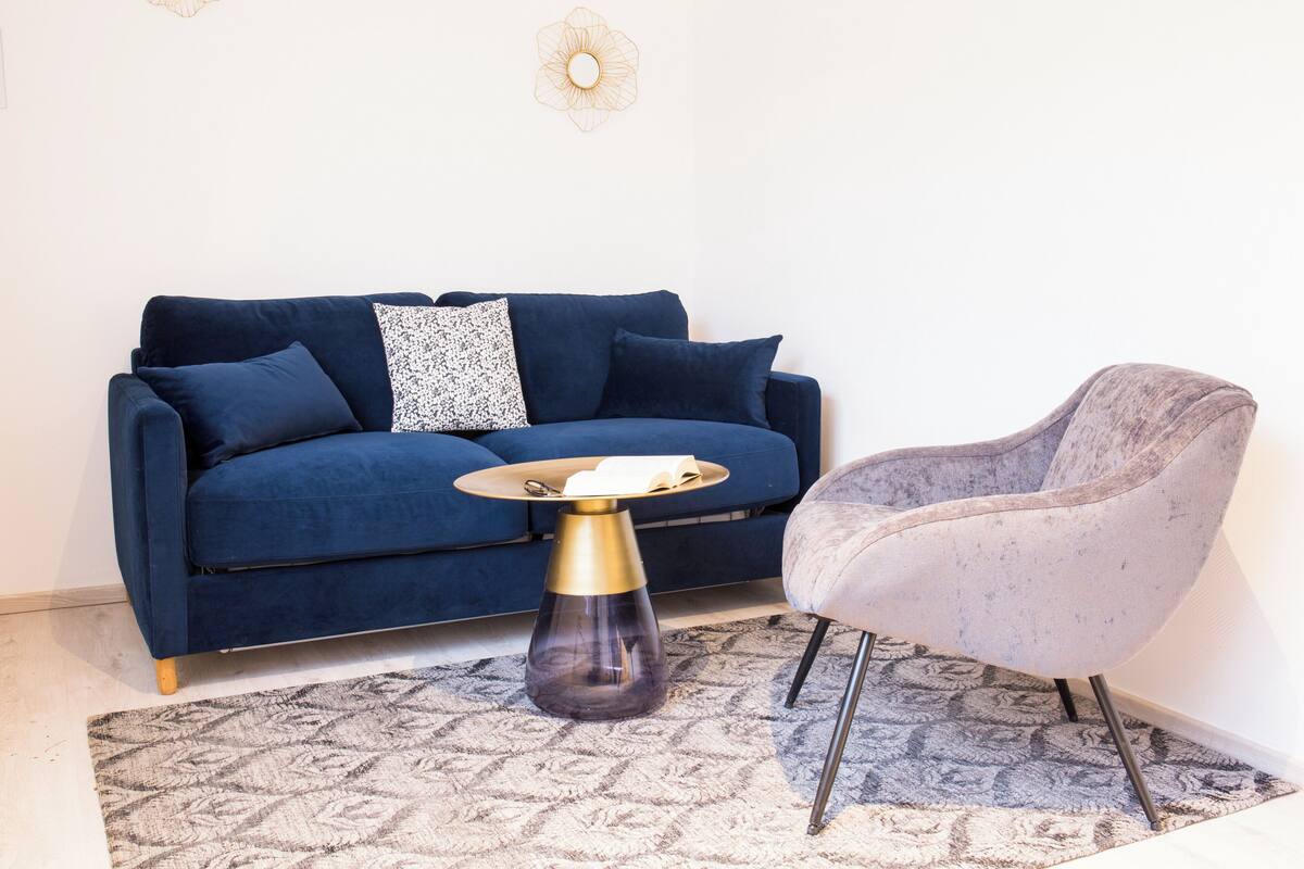 Une pièce avec un canapé bleu foncé, une table basse ronde dotée d’un pied doré, un fauteuil gris et un tapis à motifs.