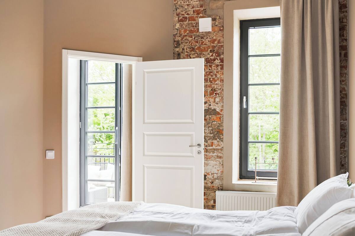 A bedroom with a brick wall, a large window with curtains, a white door, and a bed with white bedding.