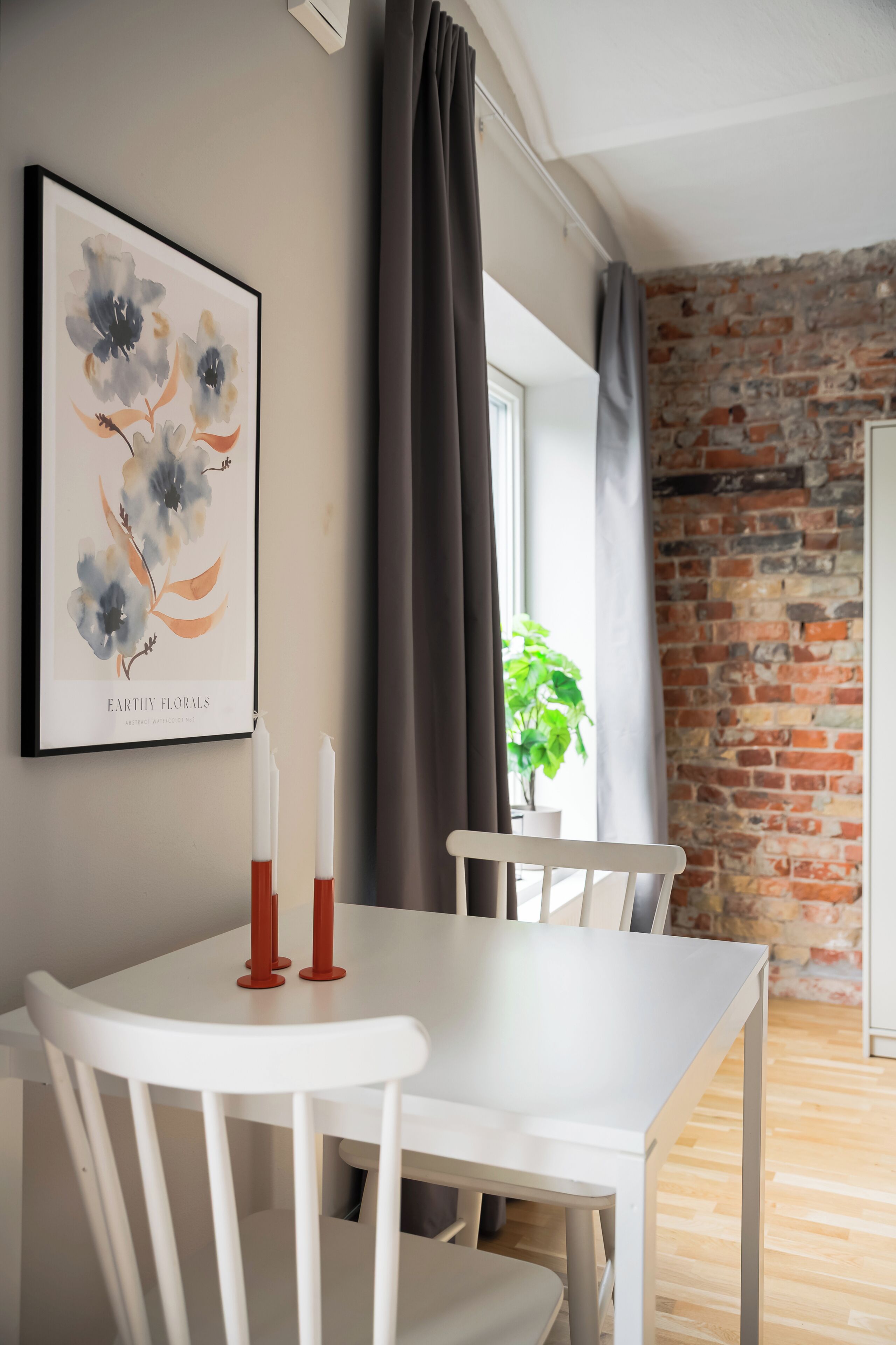 A dining area with a white table and chairs, a framed floral artwork on the wall, and a brick wall in the background.
