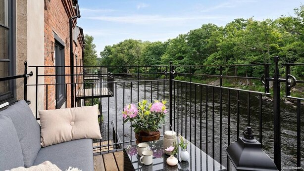 A balcony with a sofa, a table with decorative items, and a view of a river and trees. - Mörrum River  (Mörrum)