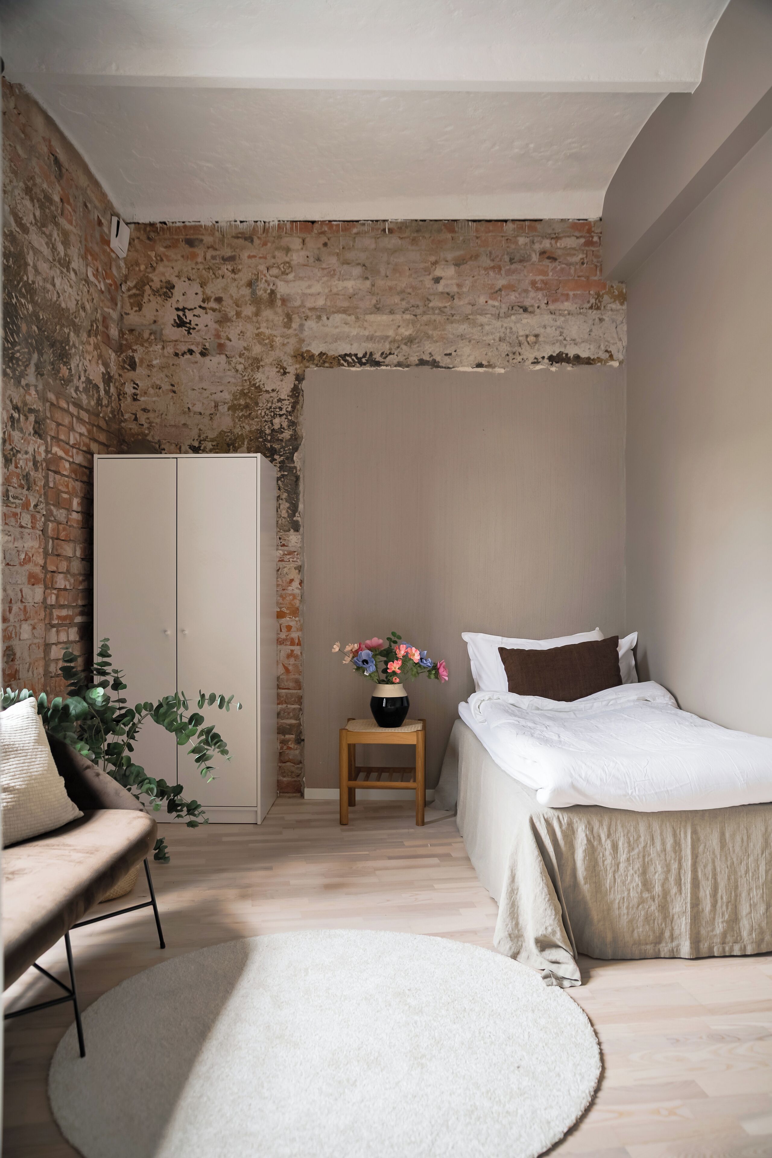 A bedroom with a brick wall, a white wardrobe, a bed with white bedding, a small wooden nightstand, and a potted plant.
