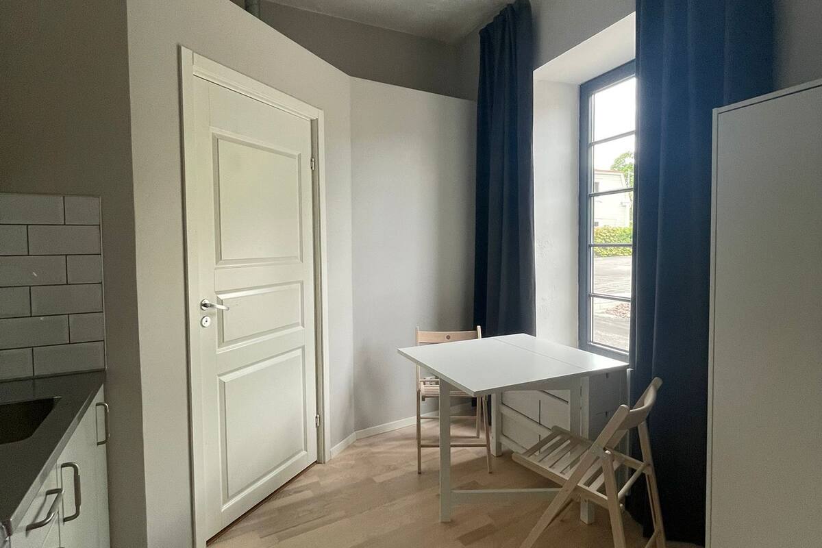 A small, well-lit room with a white door, a window with blue curtains, a white table, and a wooden chair.